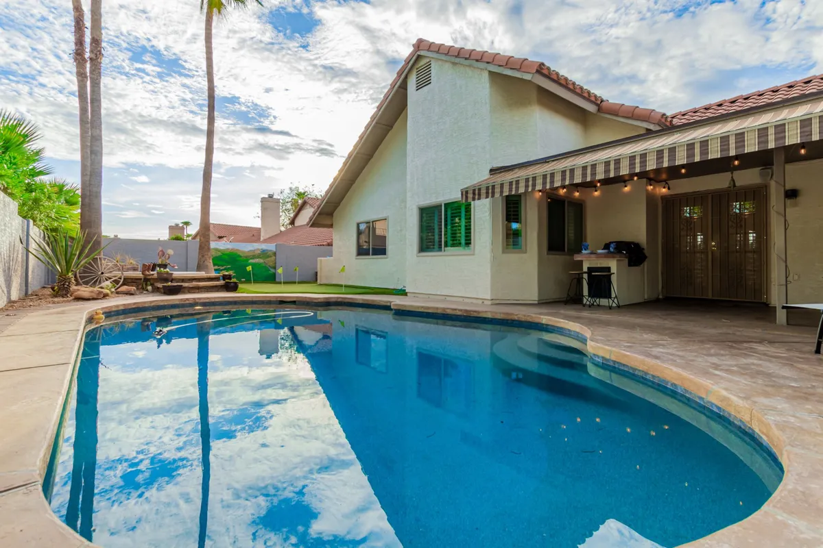 This inviting exterior image features the swimming pool, patio area, lush palm trees, and the house facade. The environment is relaxed, perfect for gathering with friends or enjoying a quiet morning before heading to nearby courses. The scene hints at the seamless blend of private retreat and easy access to golf activities.