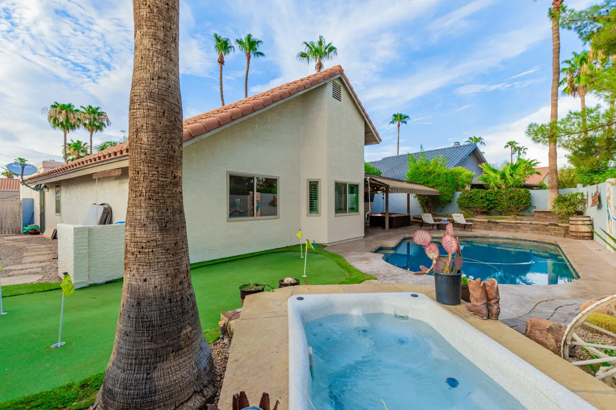 A wide-angle view of the backyard oasis, showcasing the sparkling swimming pool, an inviting hot tub, the backyard putting green, and ample outdoor seating beneath the palm trees. The scene is vibrant and designed for both relaxation and entertainment, illustrating the full breadth of resort-style amenities.