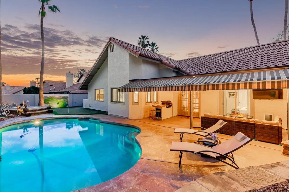 A serene backyard scene featuring a sparkling swimming pool surrounded by lounge chairs, a shaded outdoor dining area, and lush palm trees. The space feels open and inviting, perfect for both sunbathing and group gatherings. The area is secluded and has plenty of room for family activities.