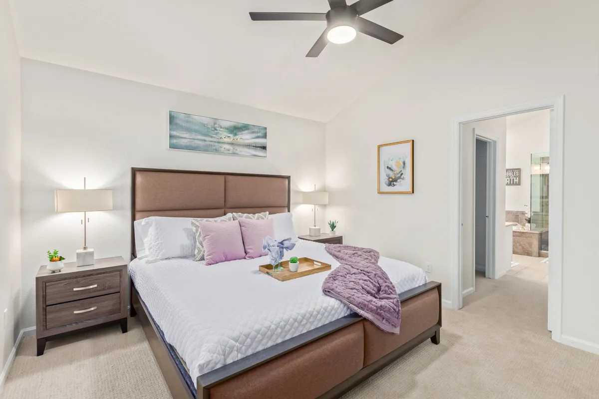 This image showcases a serene bedroom featuring a plush bed dressed in luxurious linens, decorative pillows, and nightstands with lamps. The room is styled with calming artwork and is bathed in natural light, evoking comfort and relaxation.