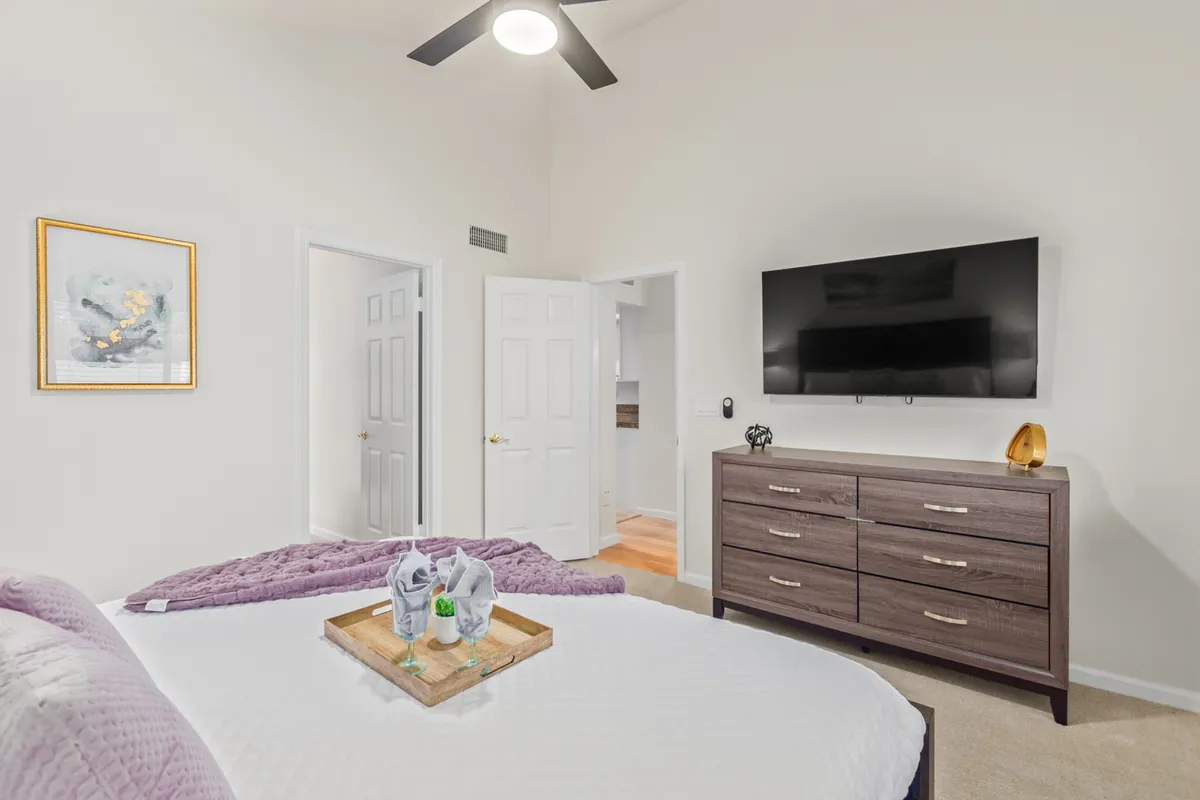 A serene bedroom scene with a bed layered in crisp white bedding, a purple throw blanket, and a dresser topped with a mounted TV. A decorative tray sits on the bed, suggesting comfort and thoughtful touches, while natural light streams into the room.
