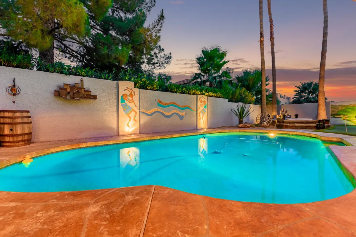 A stunning backyard scene featuring a sparkling swimming pool surrounded by palm trees and decorative wall art. The area is perfect for swimming, relaxing, or entertaining, with an inviting atmosphere that hints at both activity and relaxation. The warm sunlight and lush greenery create a resort-style vibe.